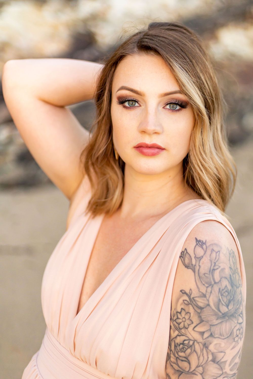 Portrait of a woman in a pink dress with blonde wavy hair and a floral tattoo on her arm.