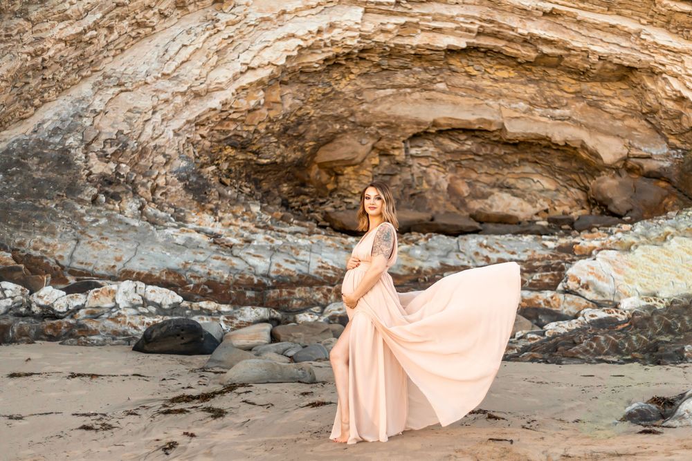 A pregnant woman in a flowing peach gown poses gracefully against dramatic coastal rock formations at sunset.