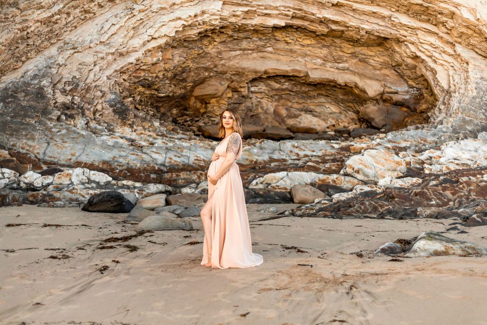 A pregnant woman in a flowing peach dress poses gracefully in front of a rocky coastal cave formation at sunset.