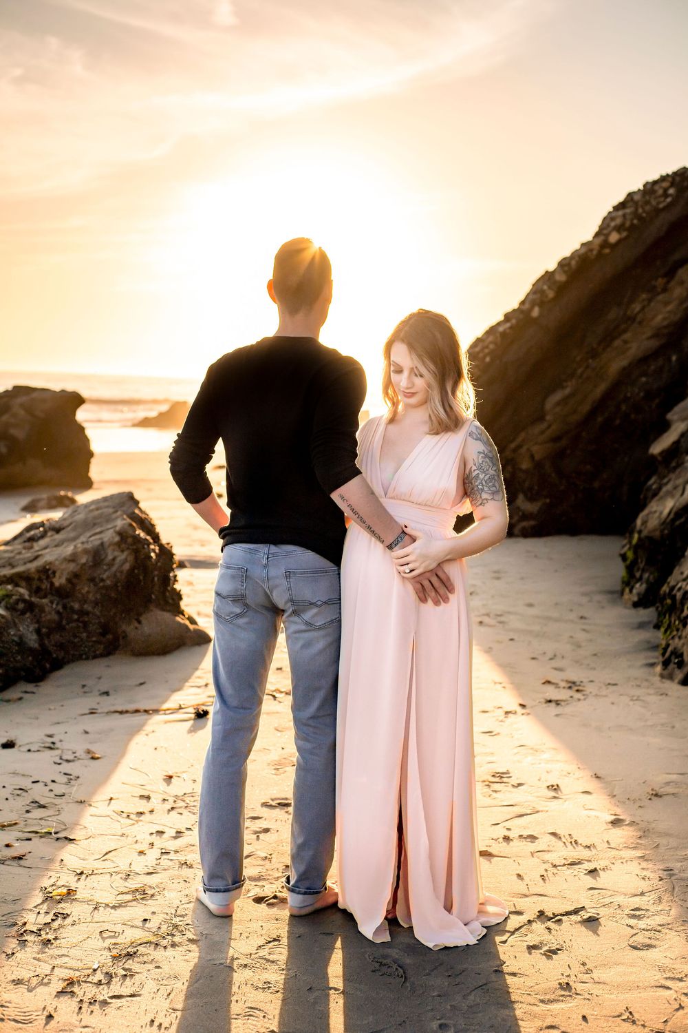 A couple stands together on a sandy beach at sunset with rocky cliffs in the background.  Woman is pregnant, facing the camera