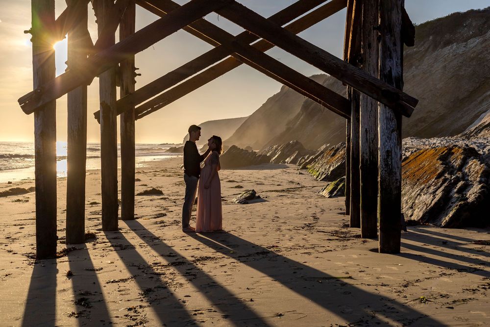 Two silhouettes embrace under wooden pier supports at sunset on a dramatic coastal beach with cliffs in the background.