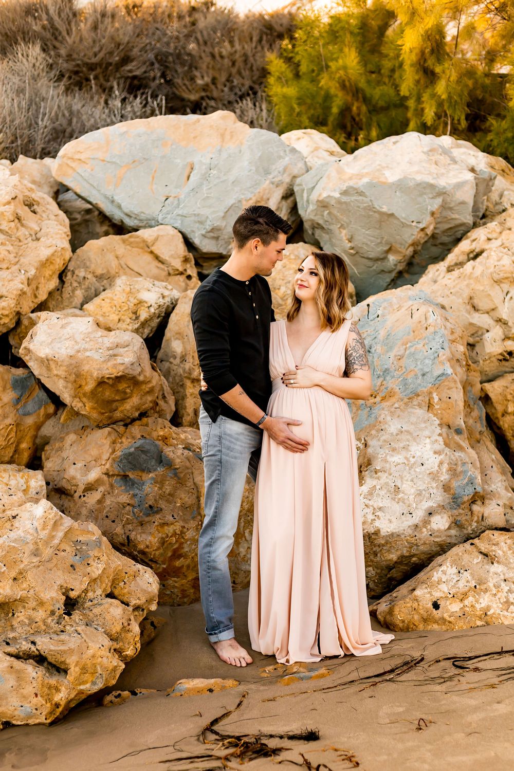 A couple shares a tender moment against large boulders at sunset, with the woman wearing a flowing pink dress is pregnant.