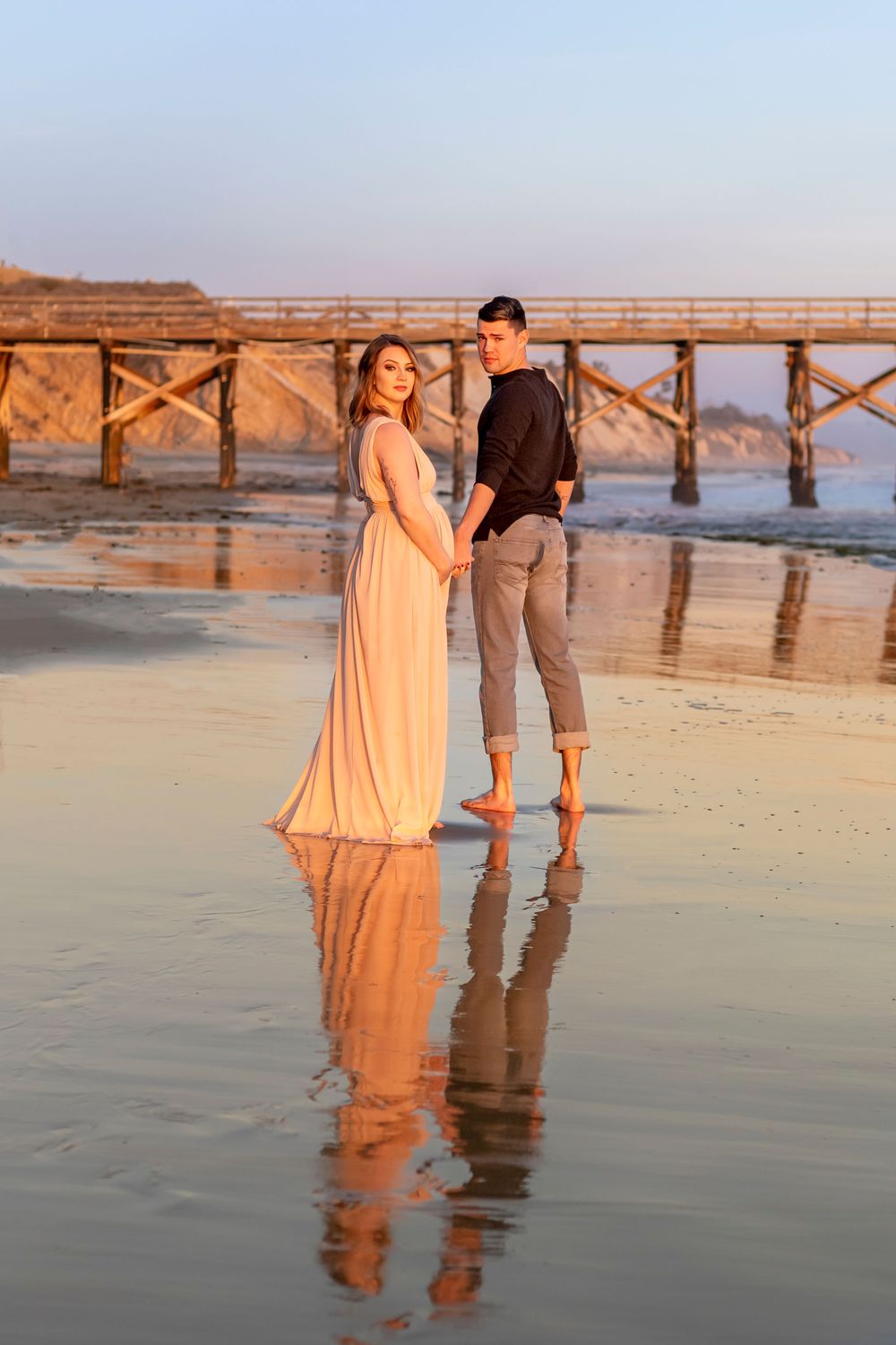 A couple stands barefoot on wet beach sand at sunset with a wooden pier in the background, creating mirror reflections.