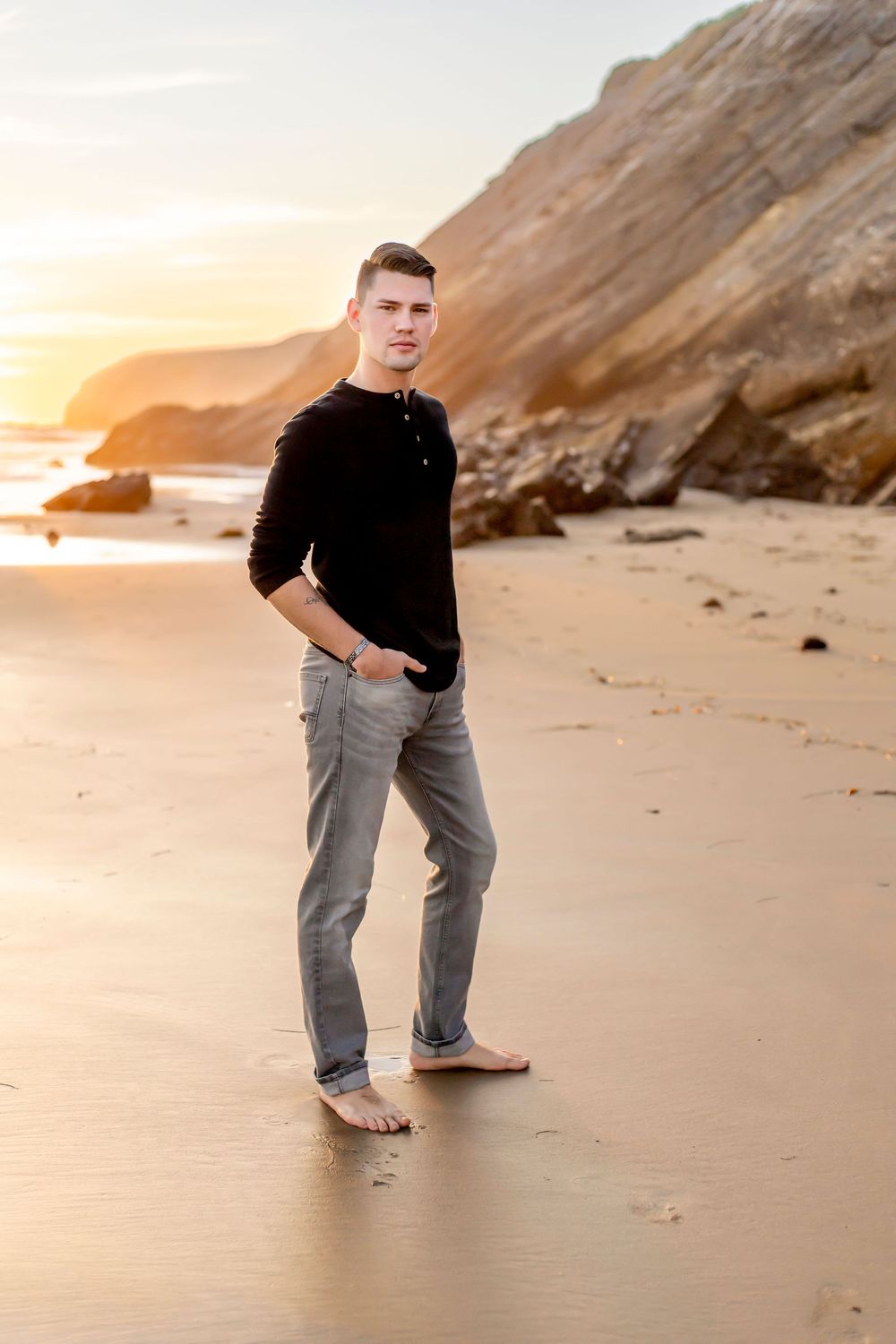 Man in black sweater and jeans standing barefoot on beach at sunset with cliffs in background.