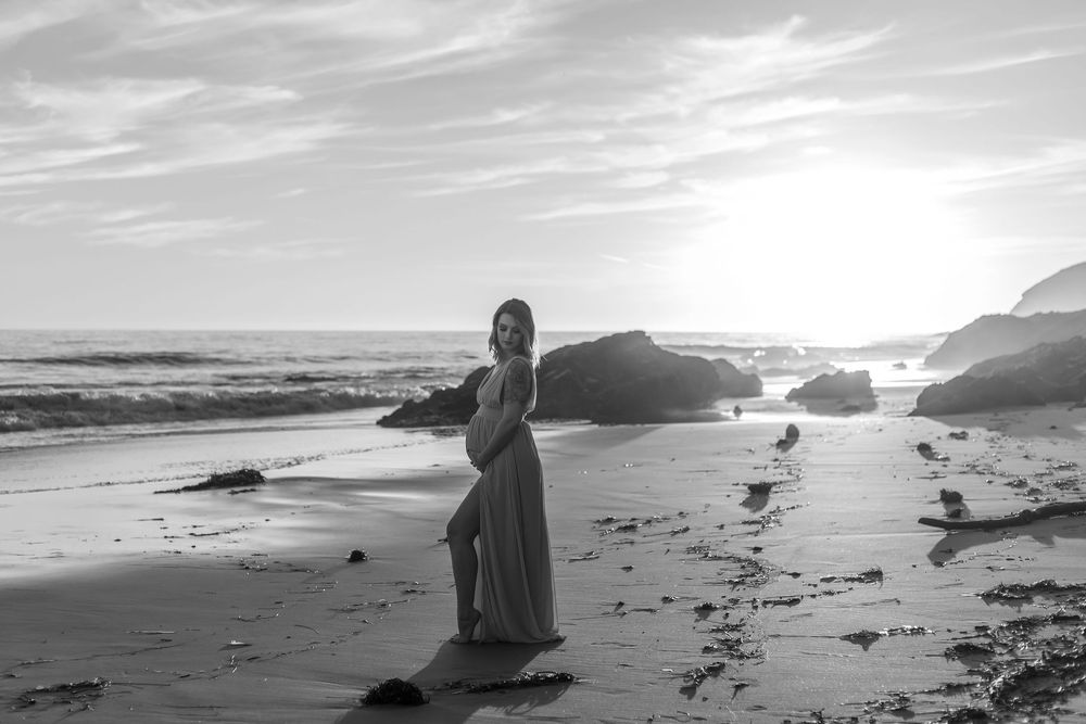 A silhouetted pregnant woman stands on a rocky beach at sunset with dramatic coastal cliffs and ocean waves in the background.