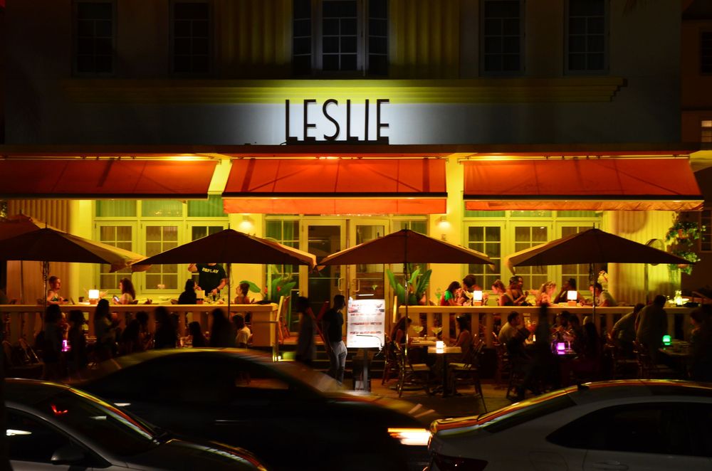 The Leslie Hotel shines with warm orange awnings and glowing lights as guests dine and socialize at packed outdoor tables.