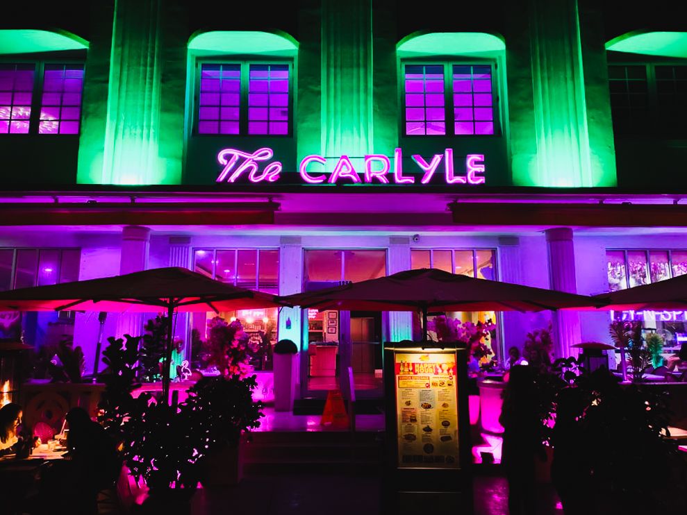 The Carlyle stands out in bold neon green, pink, and purple lighting, showcasing its iconic Art Deco architecture.