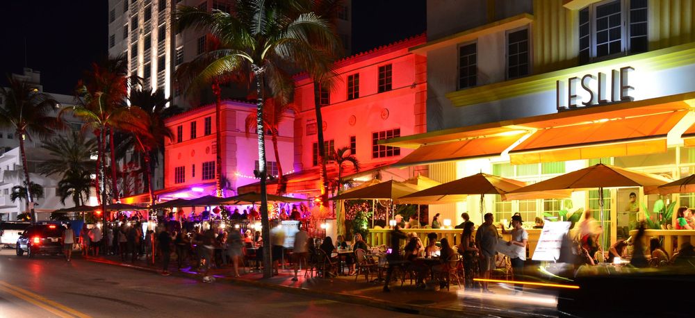 A vibrant Ocean Drive street scene comes alive with neon lights, palm trees, and bustling outdoor dining.