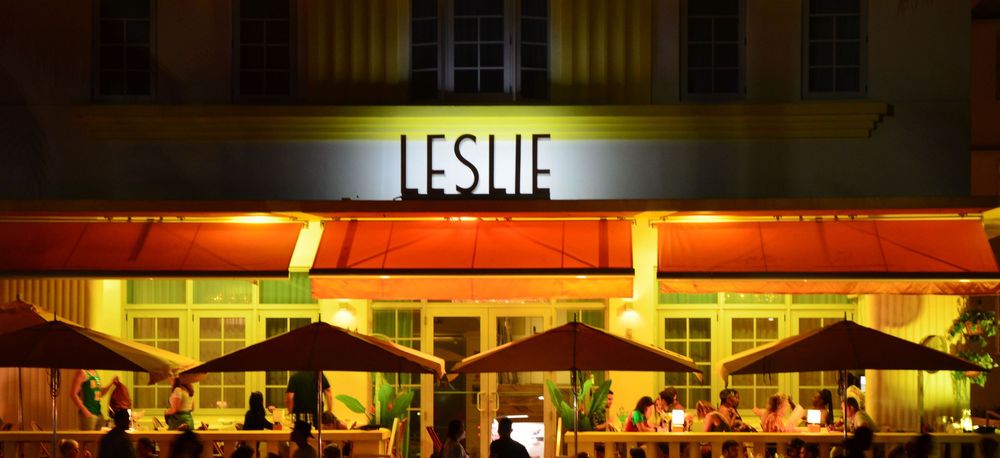 The Leslie Hotel shines with warm golden lighting, orange awnings, and lively outdoor dining that spills onto the sidewalk.
