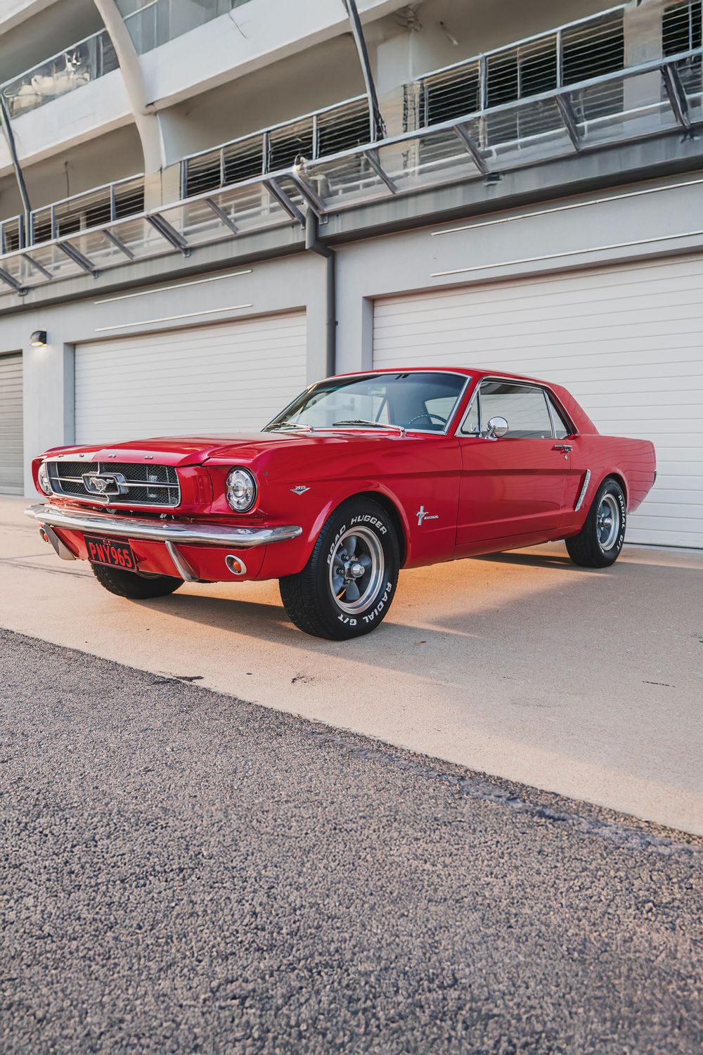 Side profile of a vintage red Ford Mustang parked against an industrial building backdrop.