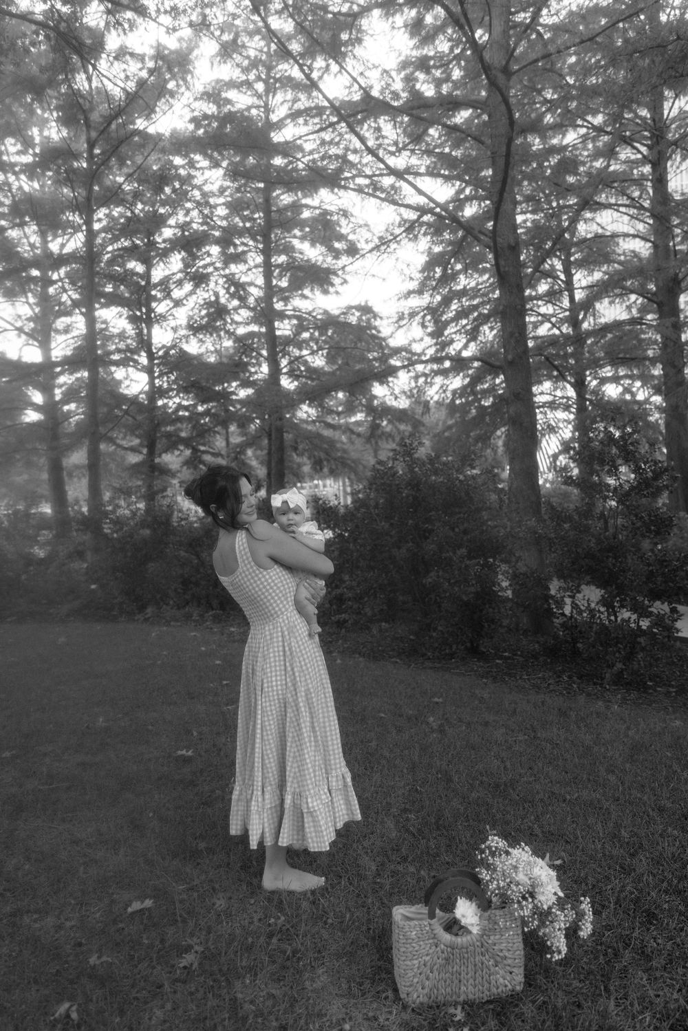 A dreamy black and white scene shows a figure in a flowing dress standing among tall trees with a basket of flowers nearby.