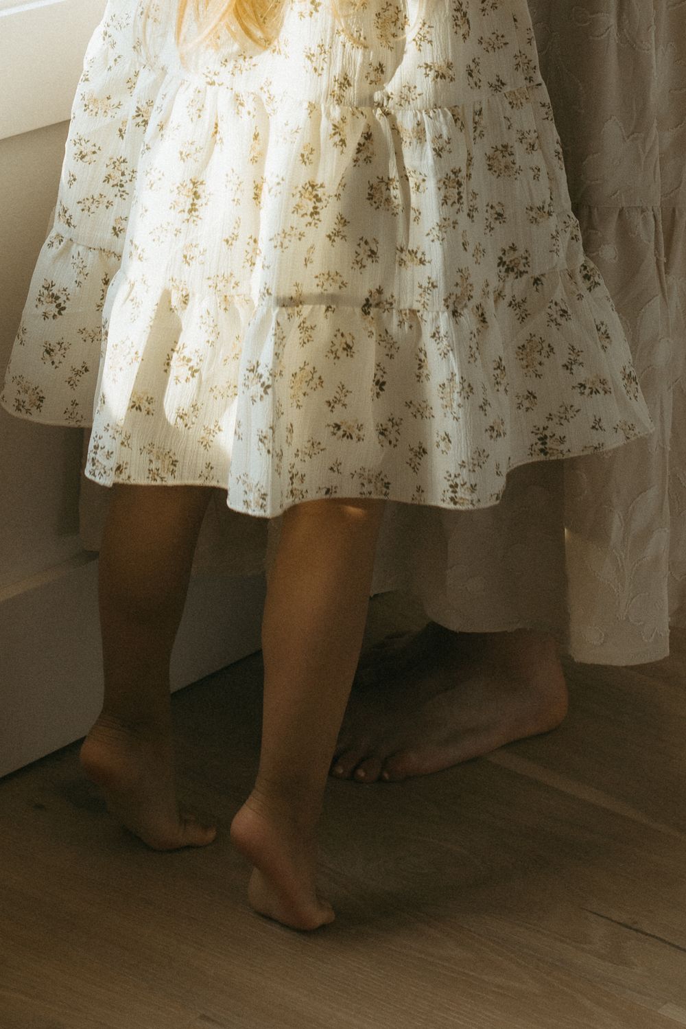 Bare feet peeking out from beneath a delicate white floral dress against a hardwood floor.