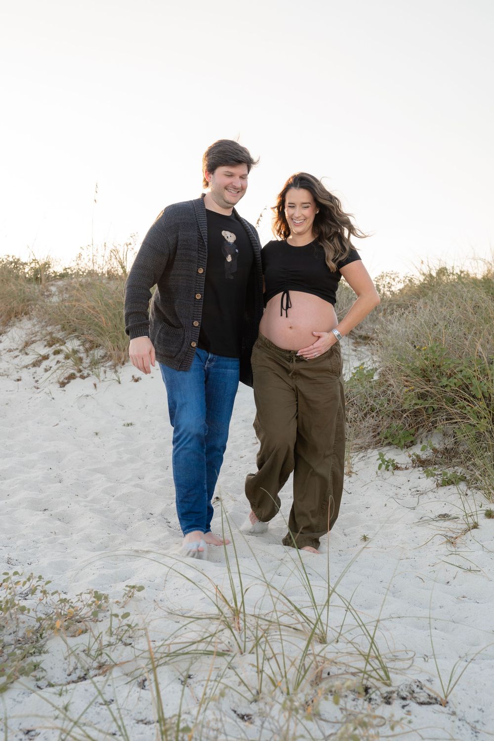 A couple walks playfully through sand dunes covered in beach grass during a coastal photoshoot.