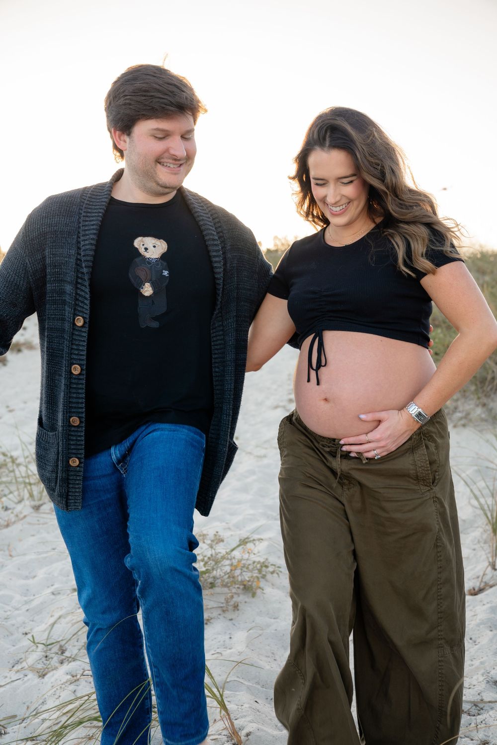 A couple smiles during their casual maternity photoshoot on a sandy beach.