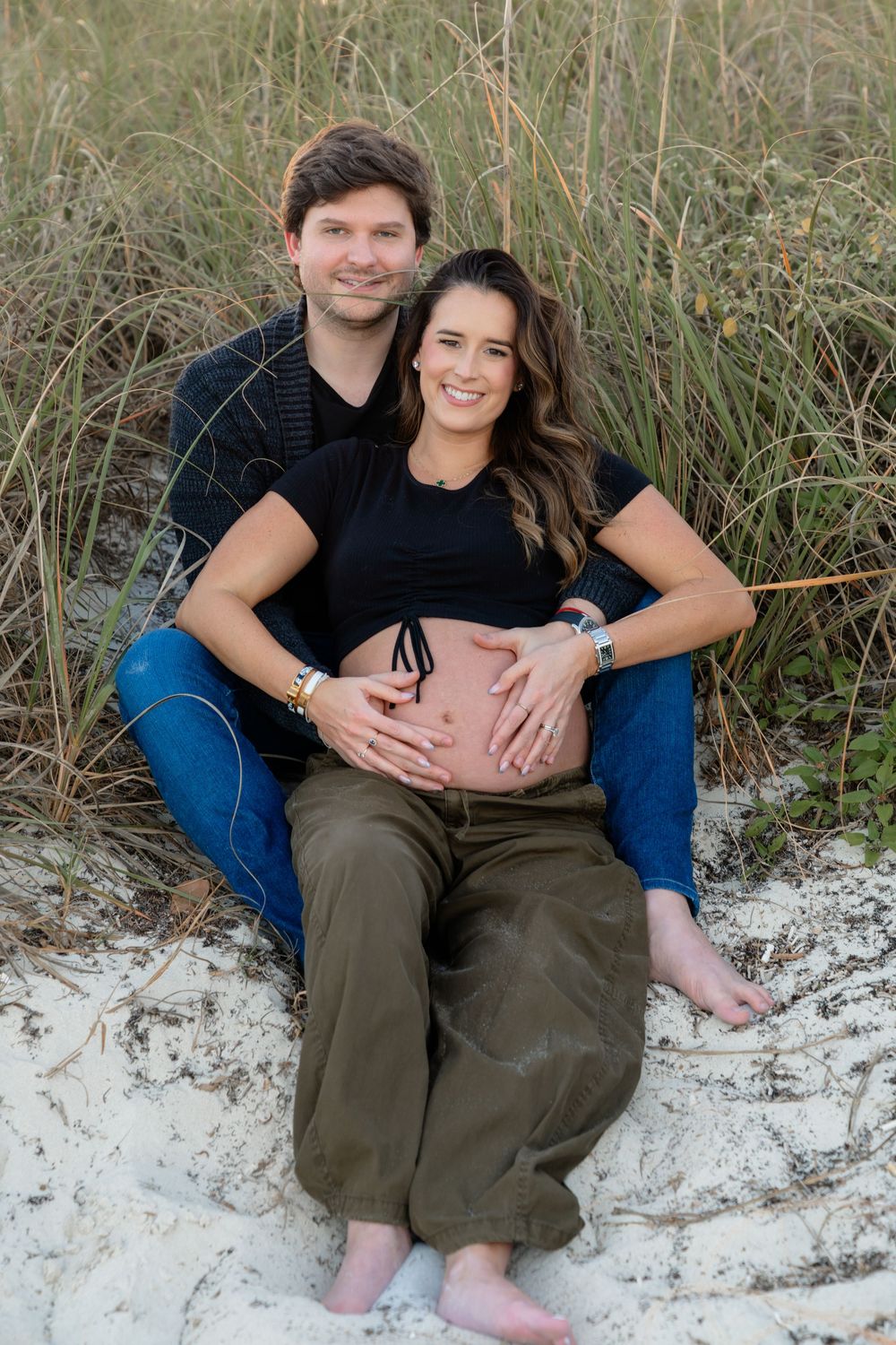 A couple sits together on white sand dunes surrounded by tall beach grass.