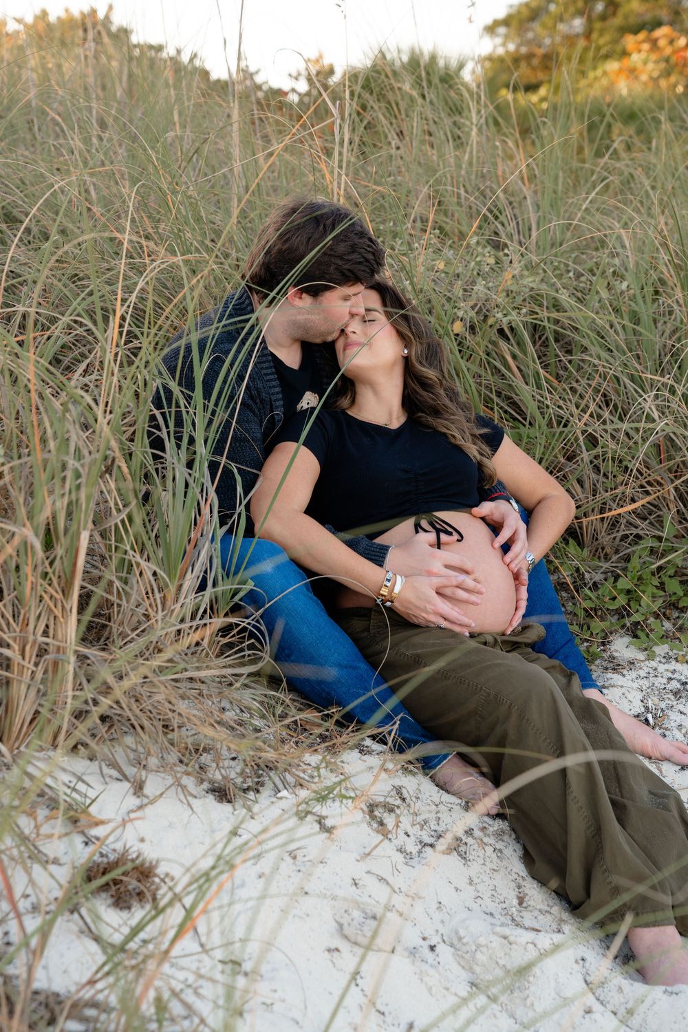 A couple relaxes in beach grass and sand during a maternity photoshoot at sunset.