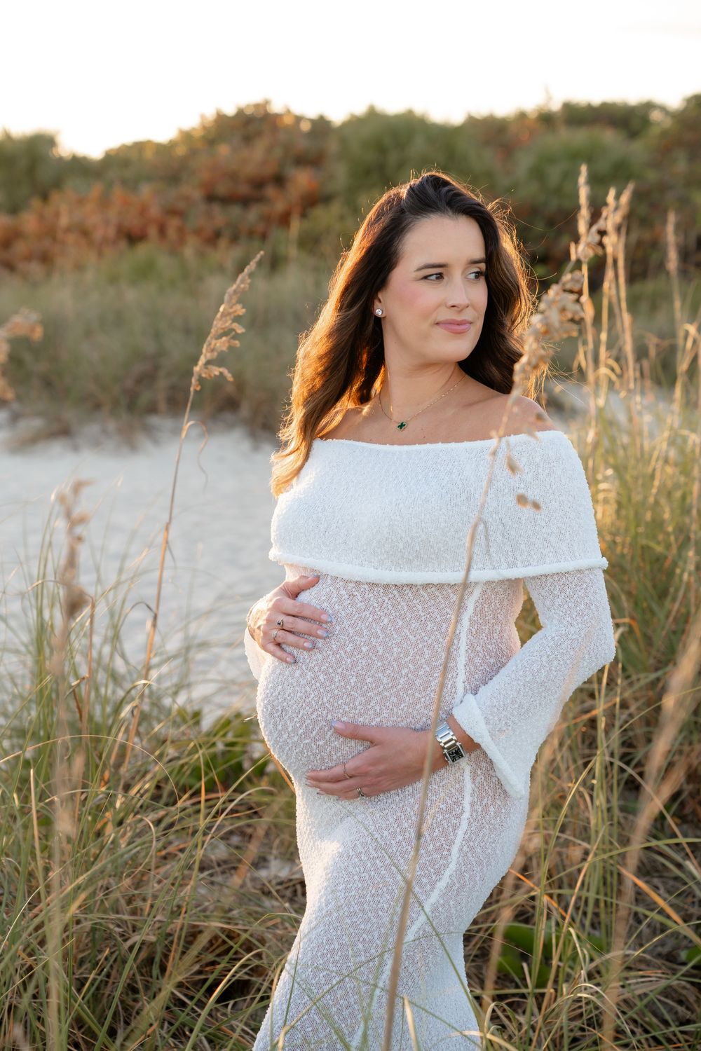 A white off-shoulder maternity dress glows in golden sunlight among beach grass during sunset.