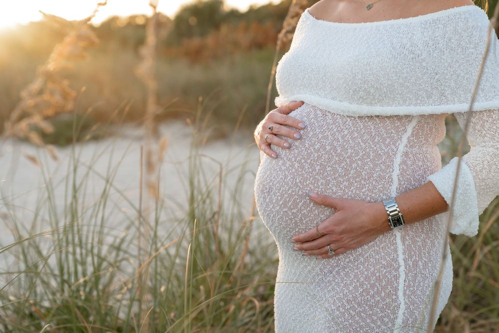A flowing white lace maternity dress is captured in warm evening light against coastal vegetation.