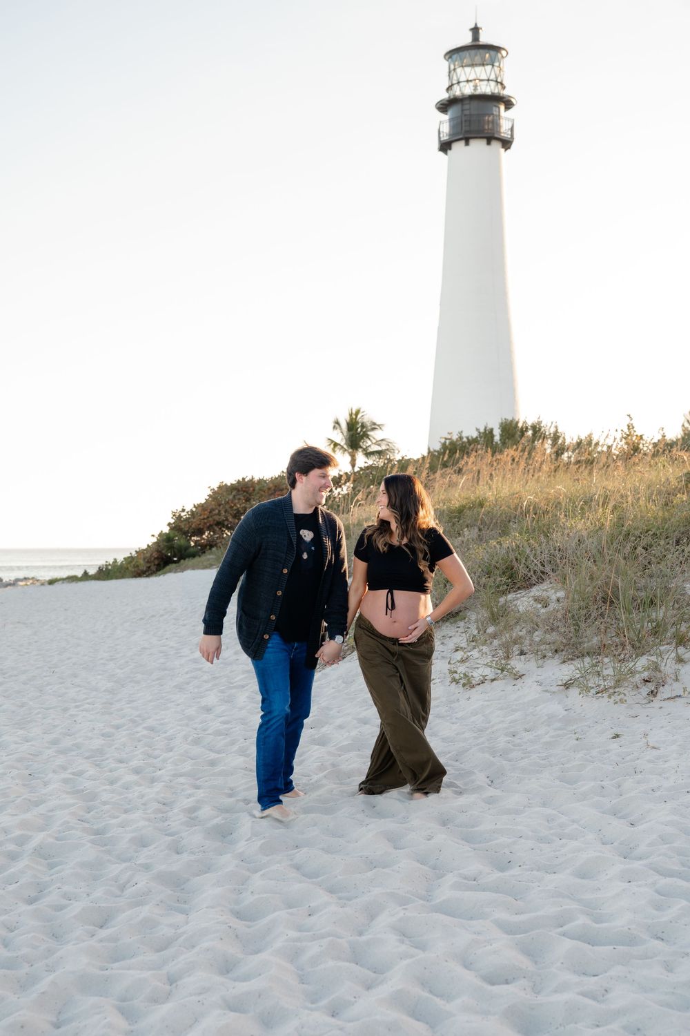A couple poses playfully near a white lighthouse on a sandy beach at sunset.
