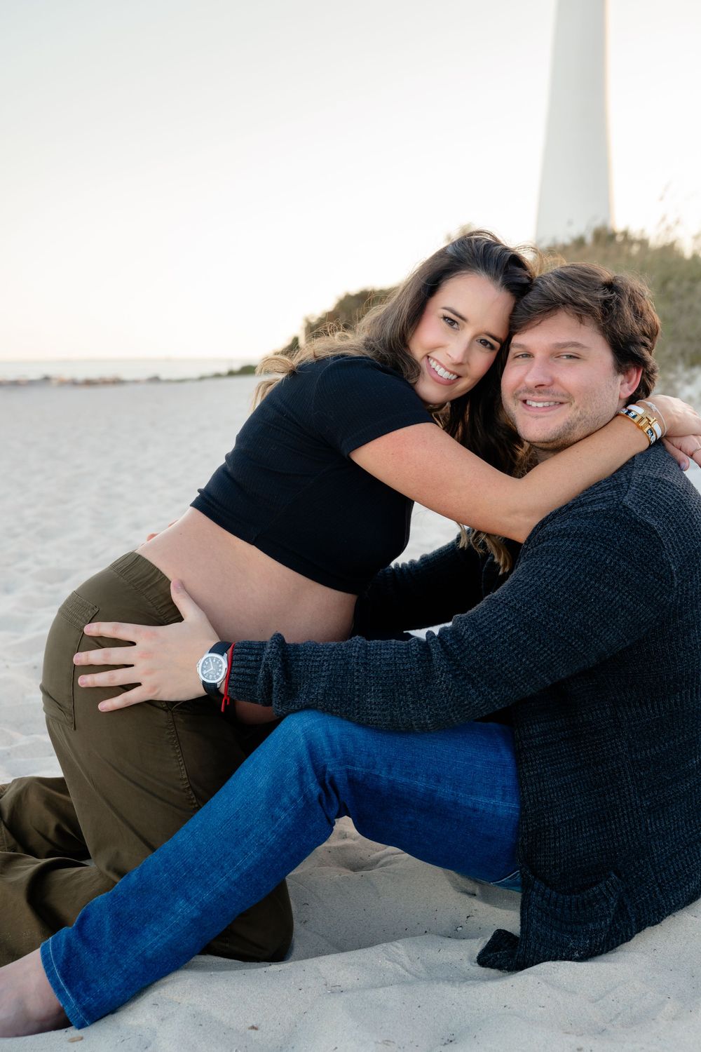 Couple embraces while sitting on a sandy beach at sunset, sharing a joyful moment together.