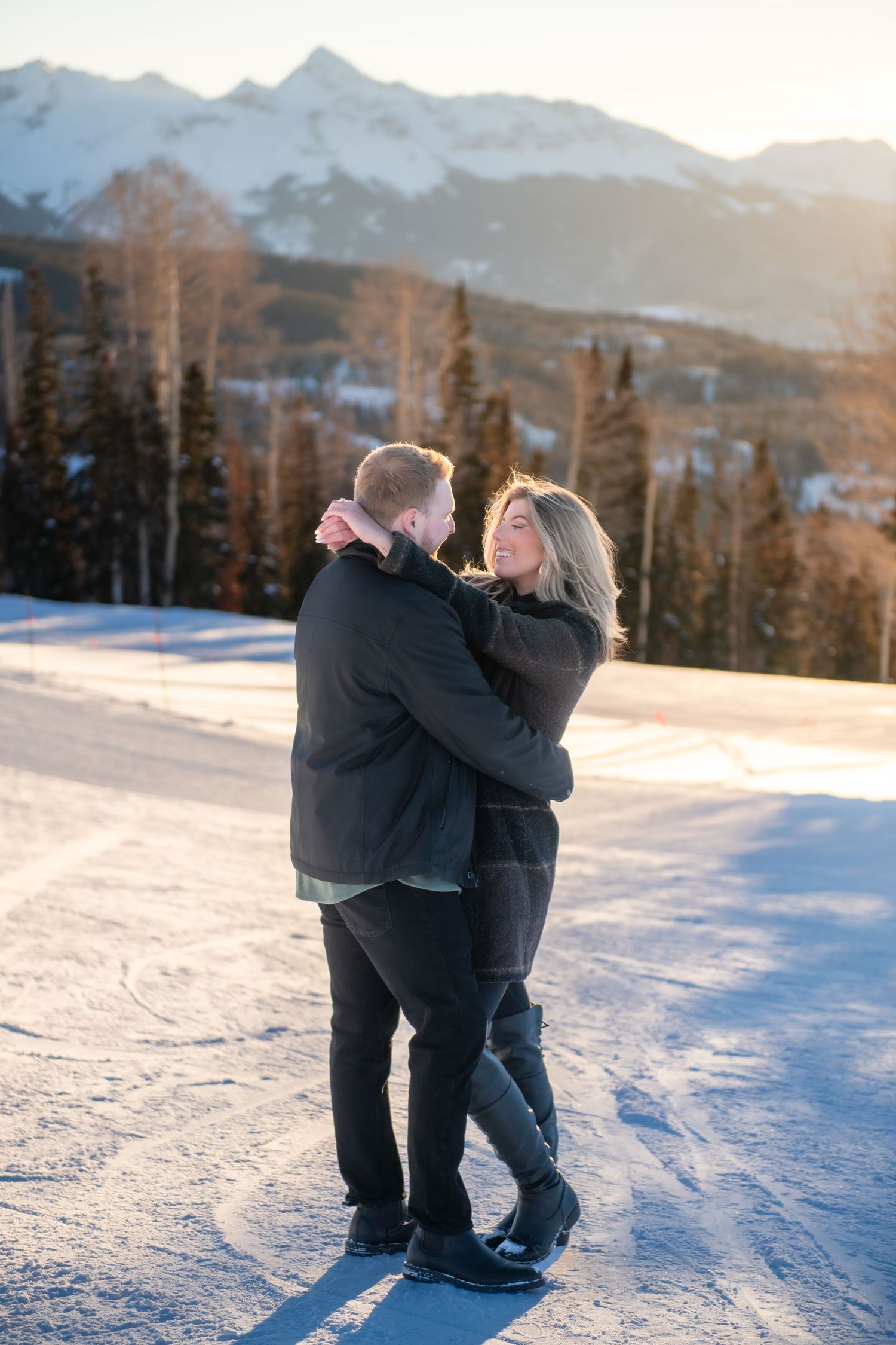 A newly engaged couple embraces on a snowy mountain slope during golden hour with evergreen trees and Mt. Wilson's peak in the background.