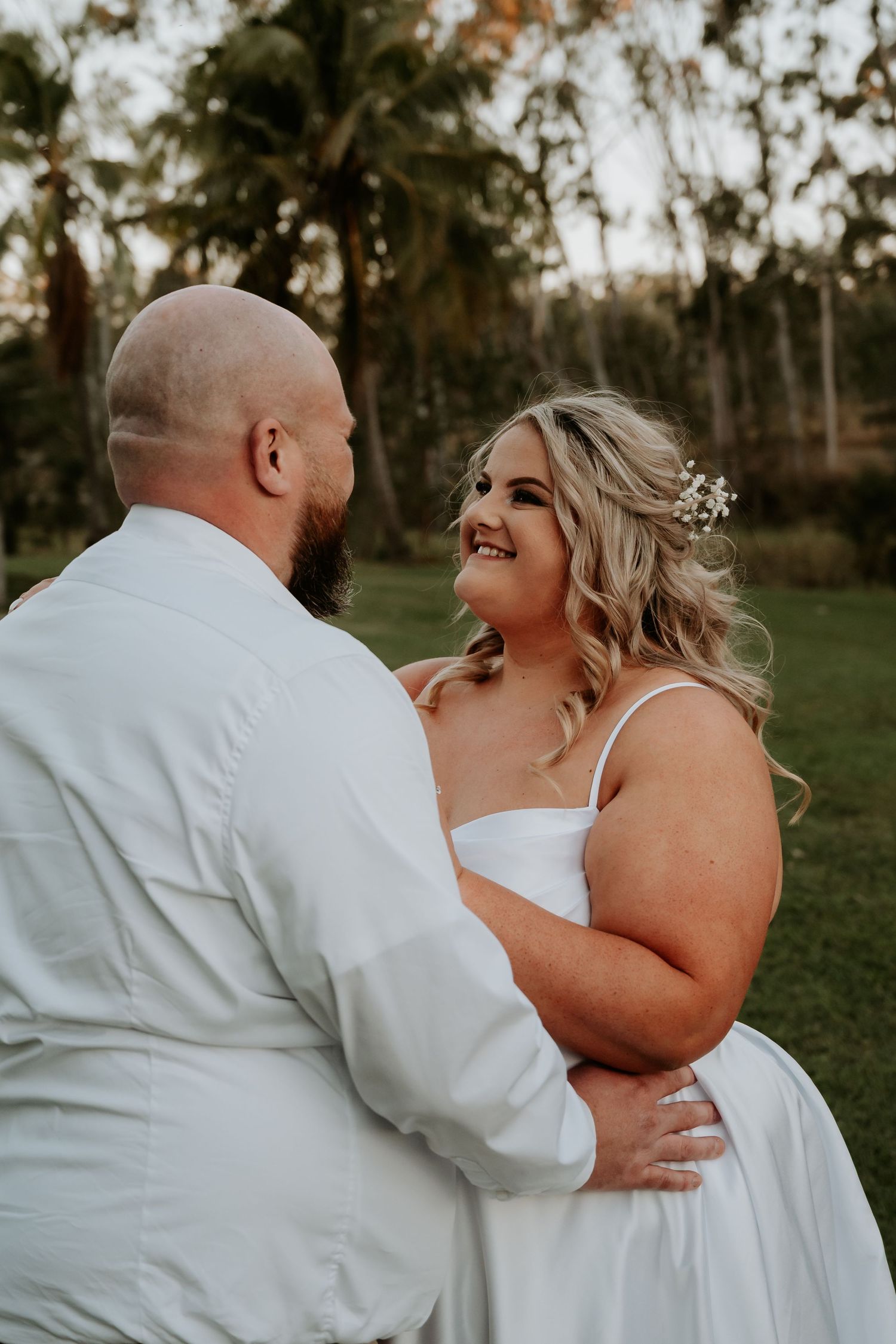 A couple in white wedding attire share candid laughing moments together in an outdoor garden setting.