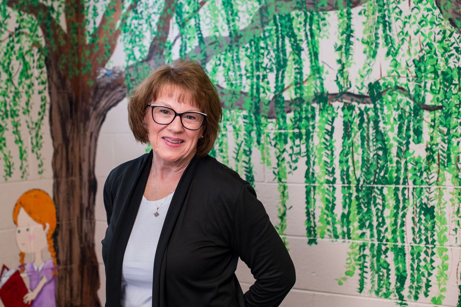 Woman smiling in front of colorful tree mural wearing black cardigan and glasses.