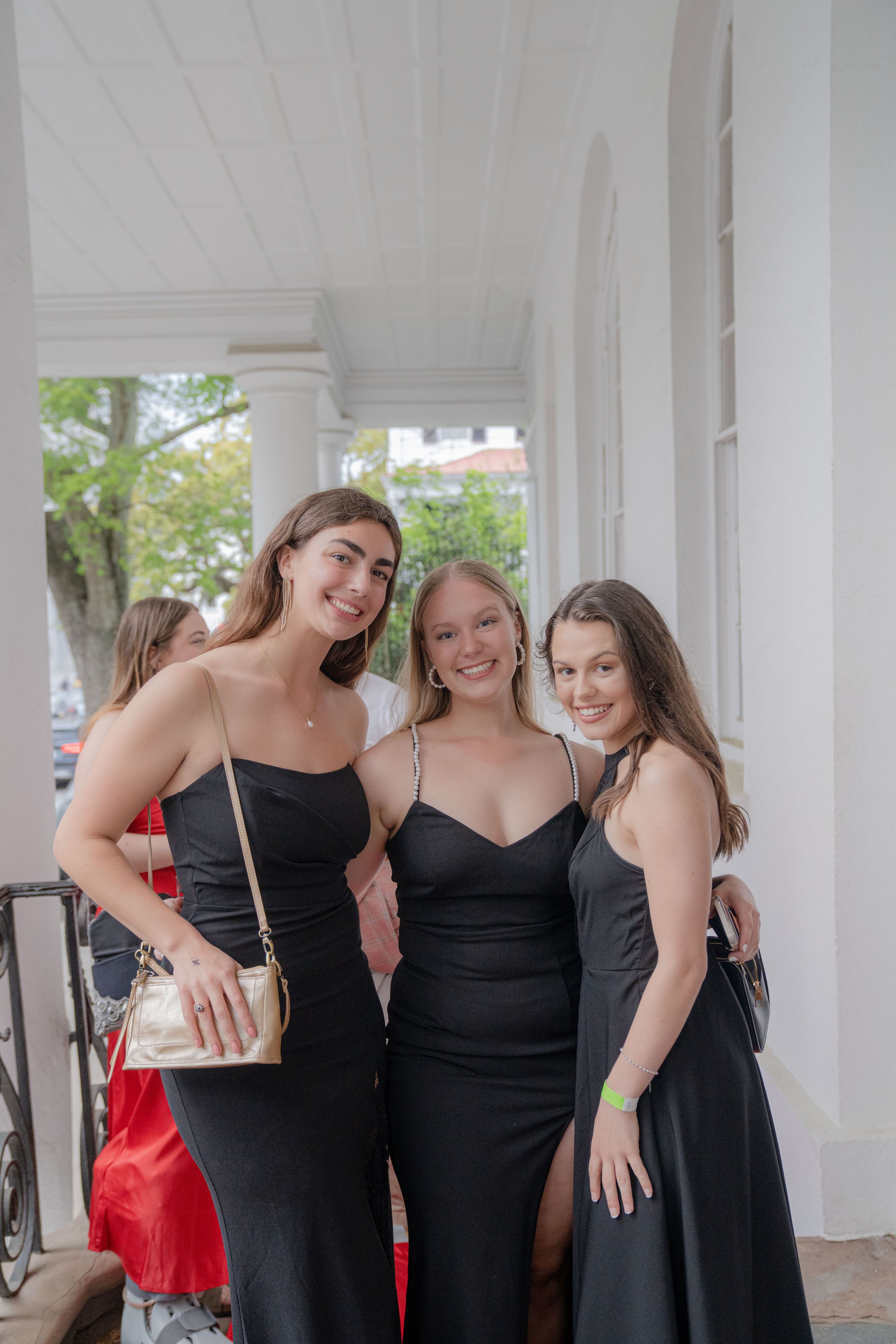 Three friends in elegant black formal dresses pose together on a covered porch during a special occasion.