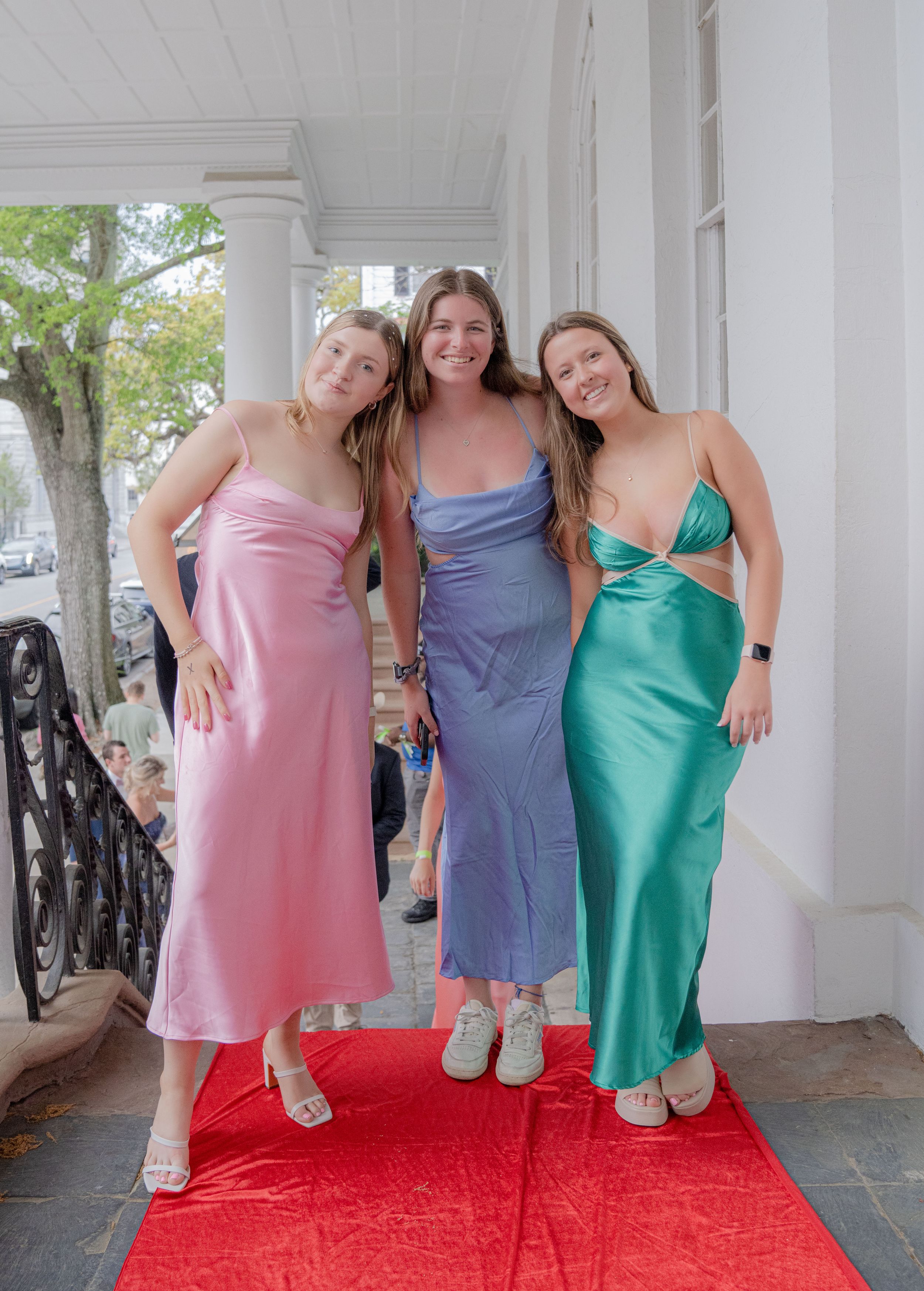 A sequence of four photos showing three friends in casual dresses posing together on a porch with a red carpet.