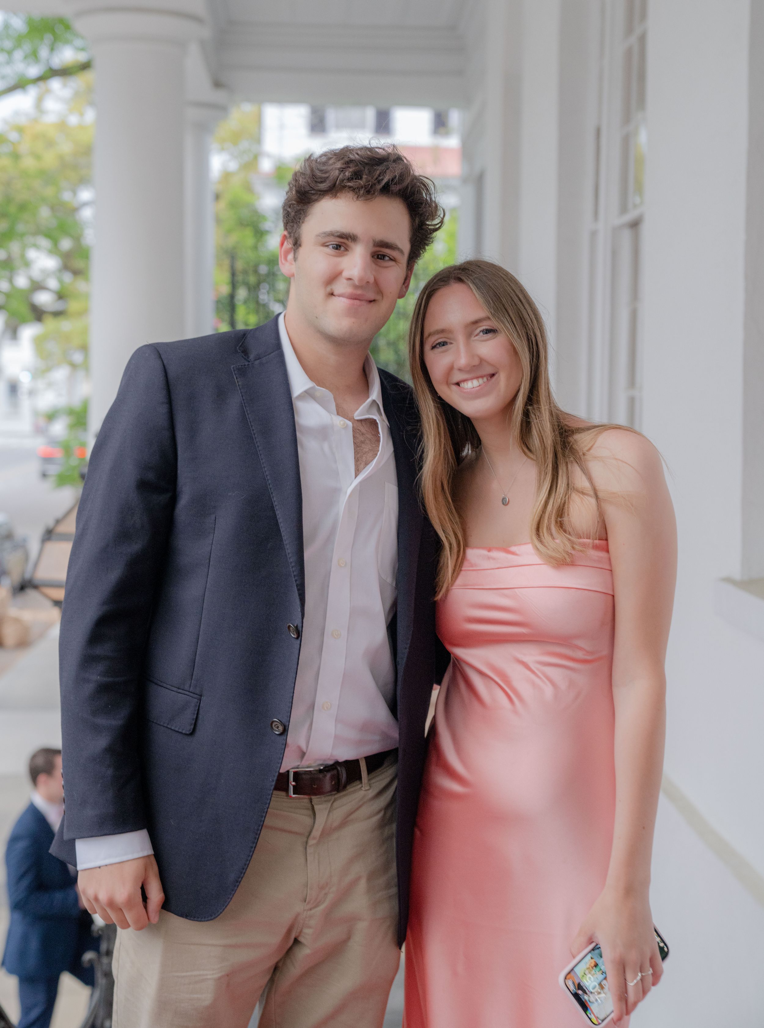 A couple poses together at an outdoor event, with one wearing a navy blazer and the other in a pink formal dress.