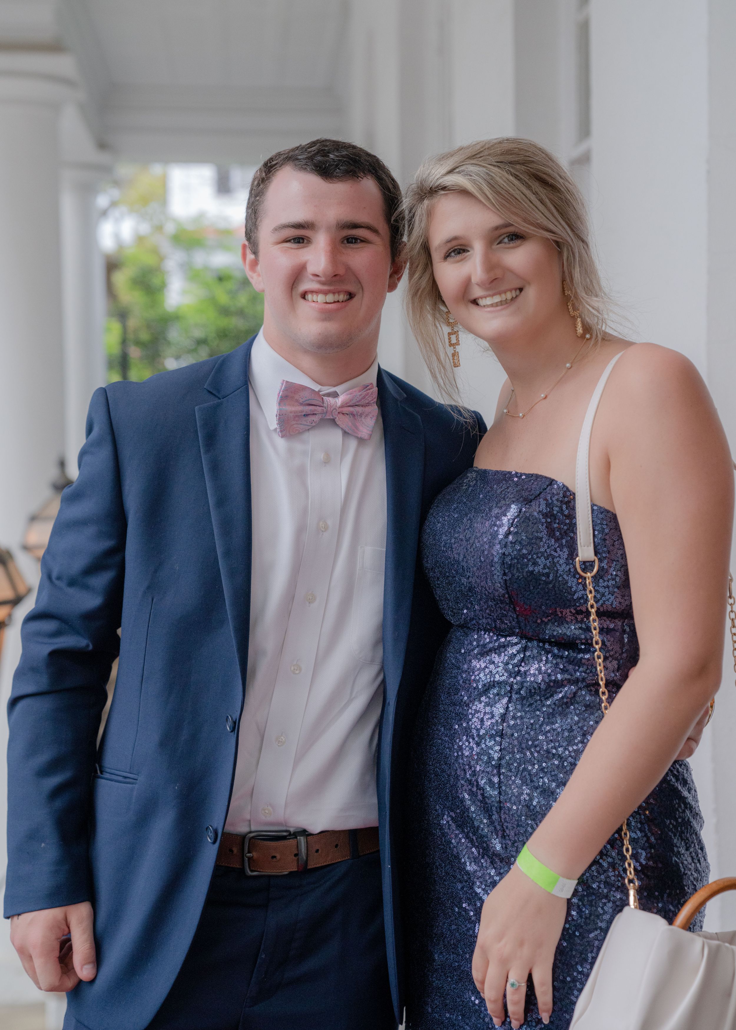 A group in formal attire poses for prom photos in a row under white columns wearing navy suits and sparkly dresses.