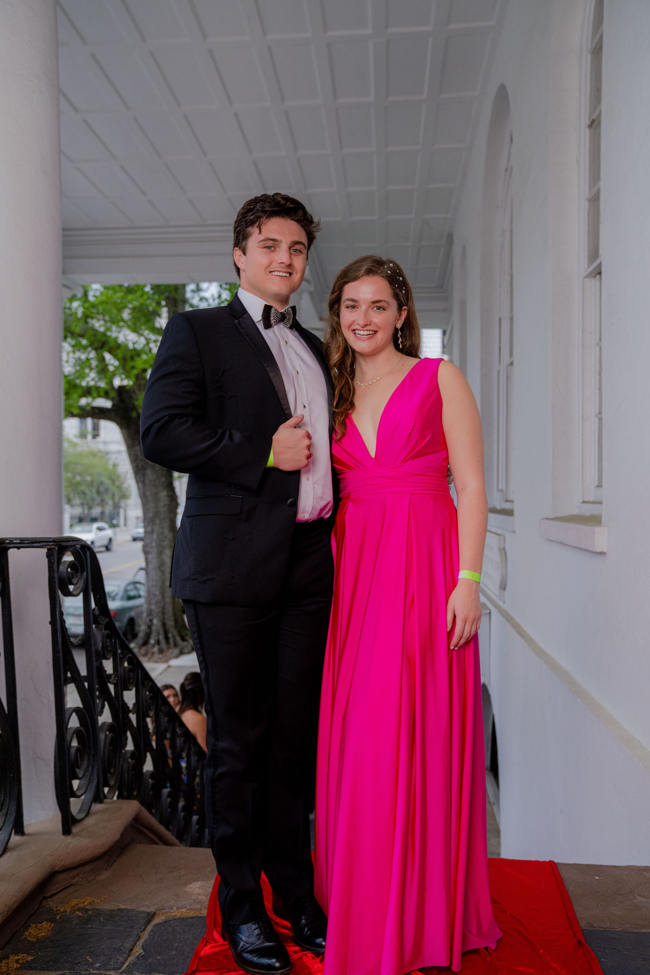 A couple poses in formal attire on a white columned porch, with the person wearing a vibrant hot pink gown.