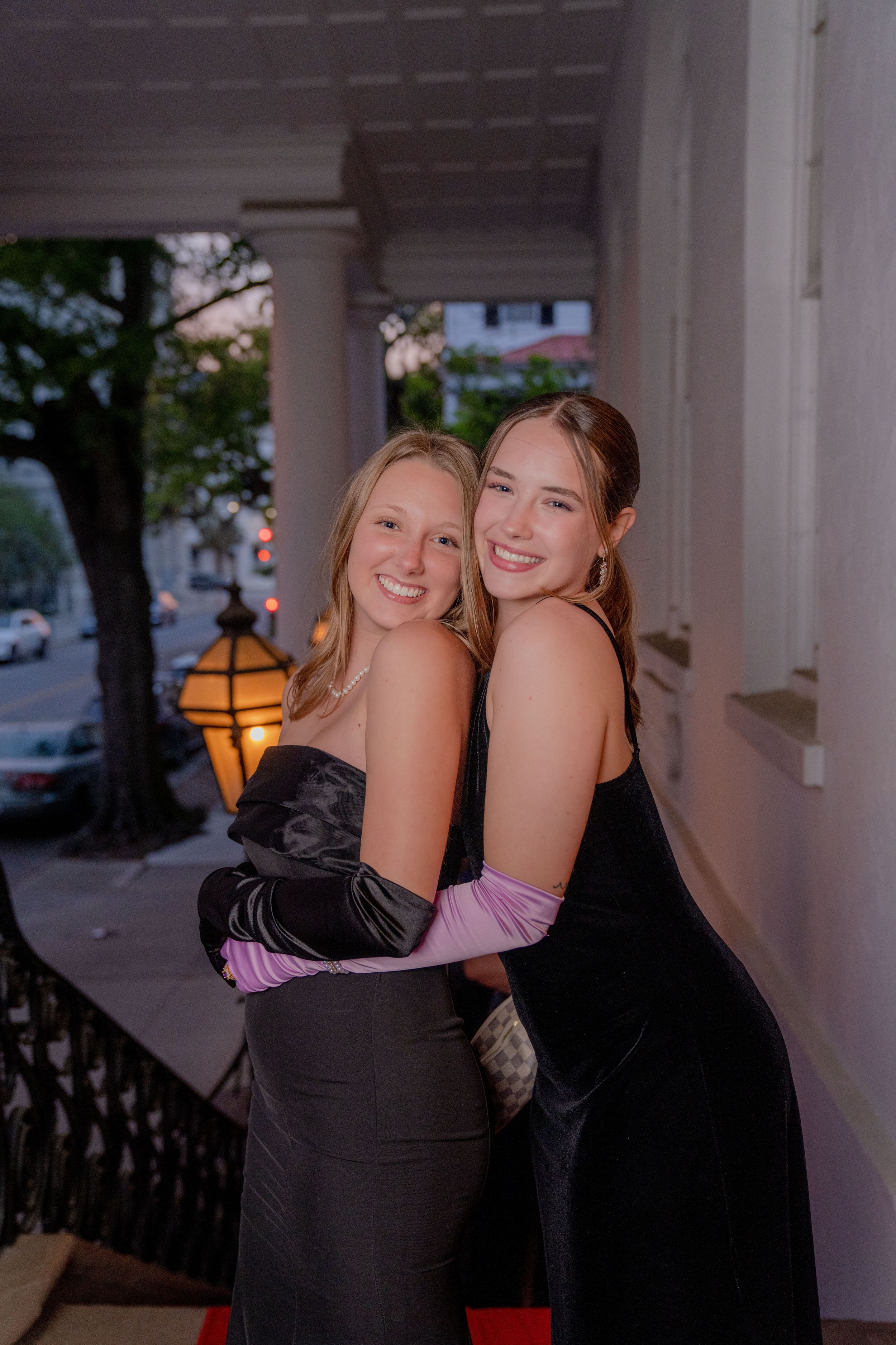 Two friends in elegant black formal dresses and gloves share a warm embrace on a porch at dusk.