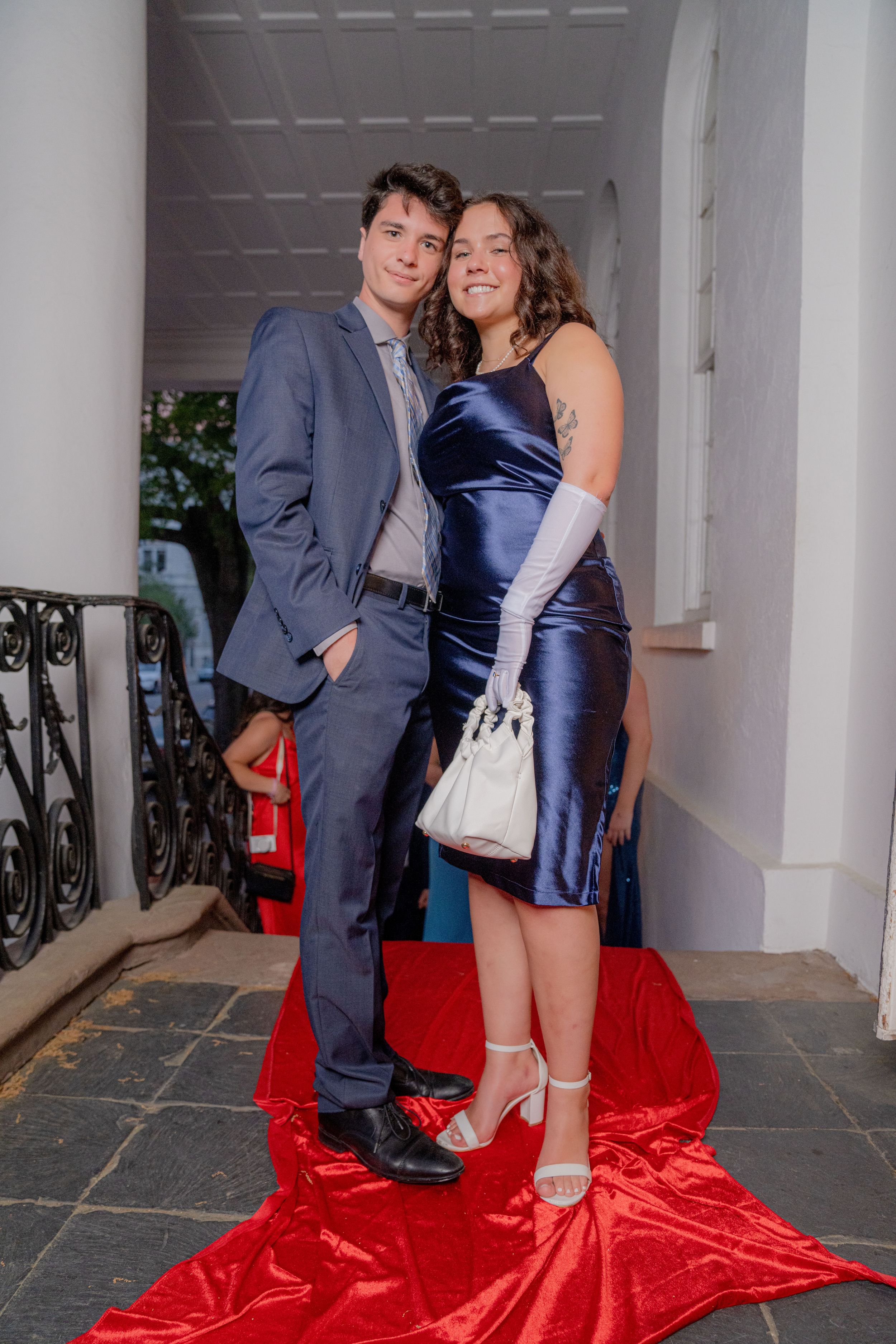 Couple poses in formal attire on red carpet in elegant hallway with white columns and iron railings.