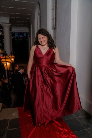 A person in a flowing burgundy ball gown twirls on a white columned porch during an evening photoshoot.