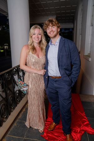 A couple poses together at a formal event, with one wearing a beige fringe dress and the other in a navy suit.