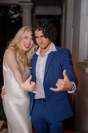 A couple in formal attire pose playfully together at an evening event on a covered porch.