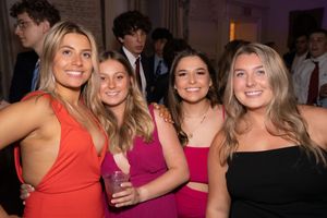 Group of friends smiling and posing together at an indoor social gathering wearing colorful cocktail dresses.