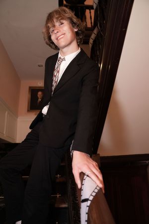 Person in a black suit poses playfully against a wall in an indoor setting.