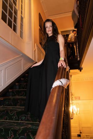 Figure in dark dress descends an elegant staircase with warm lighting and dark curtains.