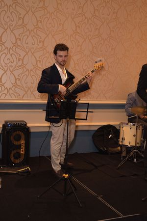 A guitarist performs on stage in a formal suit against an ornate wallpapered backdrop.