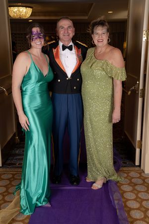 Three people in formal attire pose on a purple carpet at an elegant event, wearing green, military, and sparkly dresses.