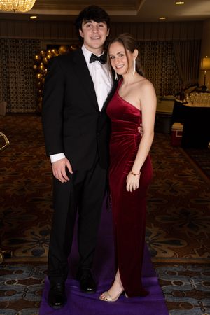 Couple in formal attire poses together at an elegant event venue with ornate carpeting and chandelier lighting.