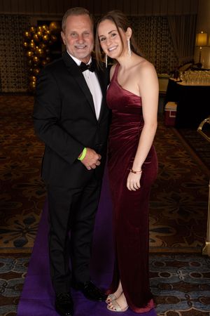 Couple in formal attire poses together at an elegant event with a burgundy velvet dress and black tuxedo.