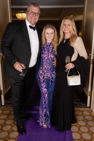 Three people in formal attire pose on a purple carpet, featuring a purple sequined gown and black tie looks.
