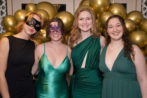 Group of women in formal attire posing together at an event with gold balloon decorations in the background.