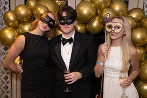 Three people in formal attire and masquerade masks pose together against a backdrop of gold balloons at an elegant party.