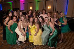 Large group poses together wearing colorful formal dresses in shades of green and yellow at an indoor event.