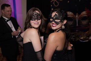 Party guests wearing black dresses and masquerade masks socialize at an elegant indoor event.