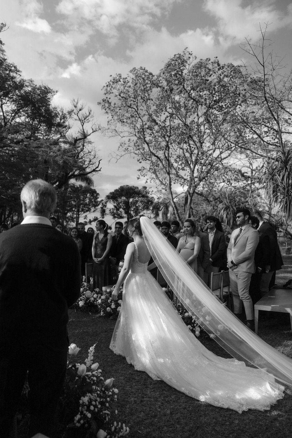A black and white outdoor wedding ceremony with a bride in a flowing gown with long veil surrounded by guests.
